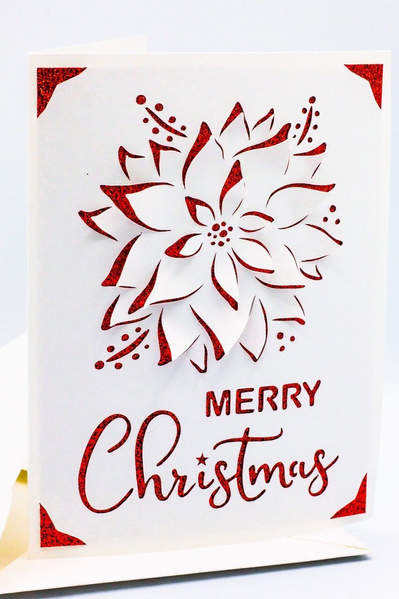 May include: A white card with a red glitter border and a red glitter cutout of a poinsettia flower. The text "Merry Christmas" is written in red glitter below the flower.