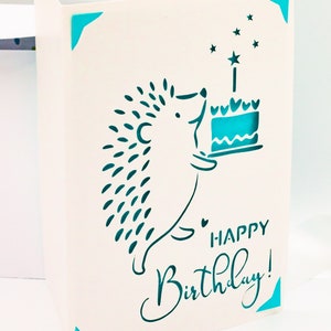 May include: A white card with a blue border and a hedgehog carrying a birthday cake with a lit candle. The card says "Happy Birthday!" in blue cursive writing.