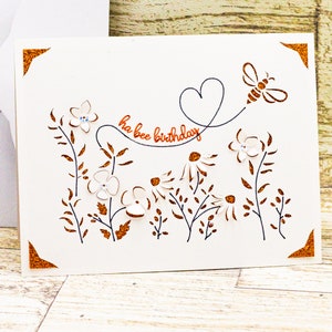 May include: A white card with a brown border and a cut-out design of flowers, leaves, and a bee. The text "ha bee birthday" is written in orange on the card.