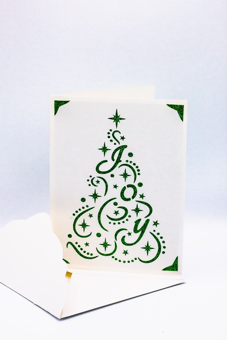 May include: A white card with a green glitter design of a Christmas tree with the word "Joy" inside. The tree is decorated with stars and swirls.