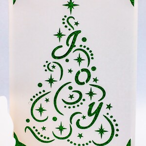 May include: A white card with a green glitter design of a Christmas tree with the word "Joy" inside. The tree is decorated with stars and crescent moons.