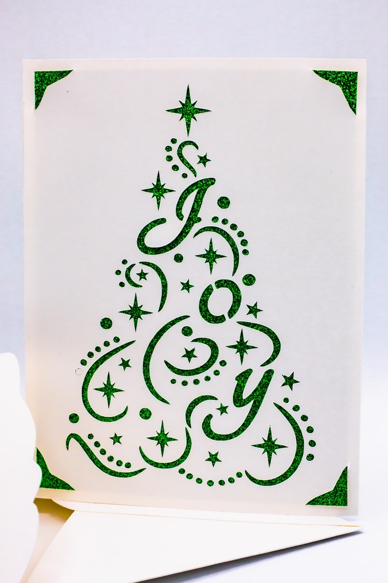 May include: A white card with a green glitter design of a Christmas tree with the word "Joy" inside. The tree is decorated with stars and crescent moons.