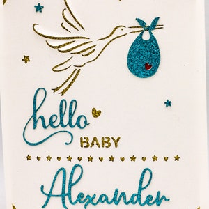 May include: A white card with a gold glitter border and a blue glitter stork carrying a blue glitter baby bag. The card says "hello baby Alexander" in blue glitter.