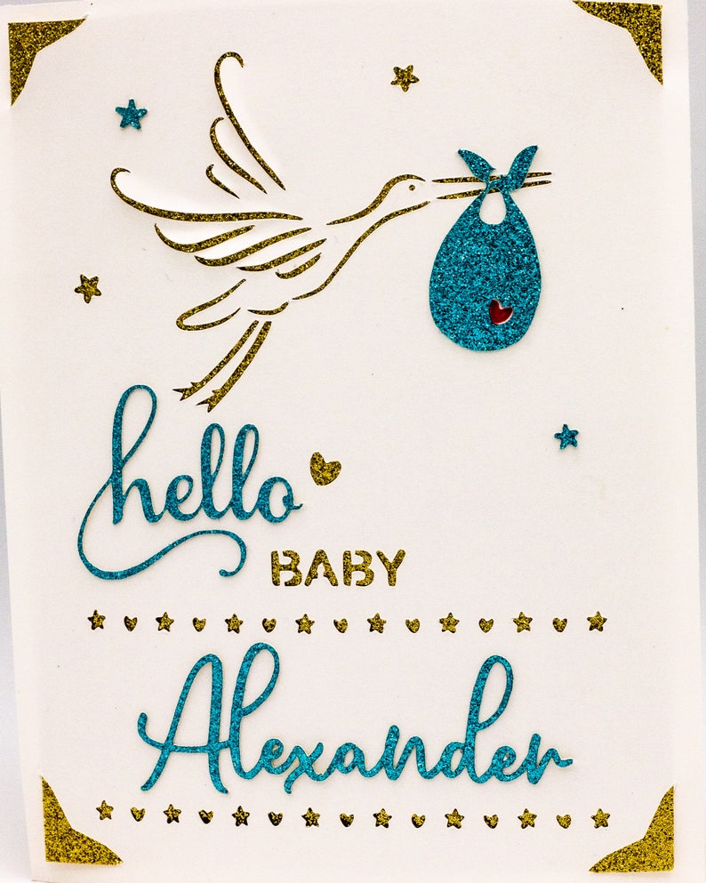 May include: A white card with a gold glitter border and a blue glitter stork carrying a blue glitter baby bag. The card says "hello baby Alexander" in blue glitter.