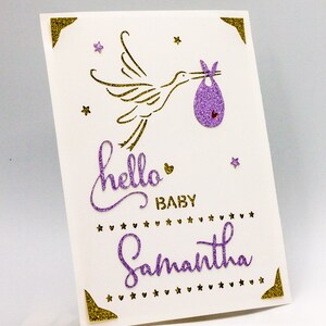 May include: A purple and gold glitter card featuring a stork carrying a baby in a purple and gold glitter baby bib. The card reads "hello baby Samantha" with gold glitter stars and hearts.