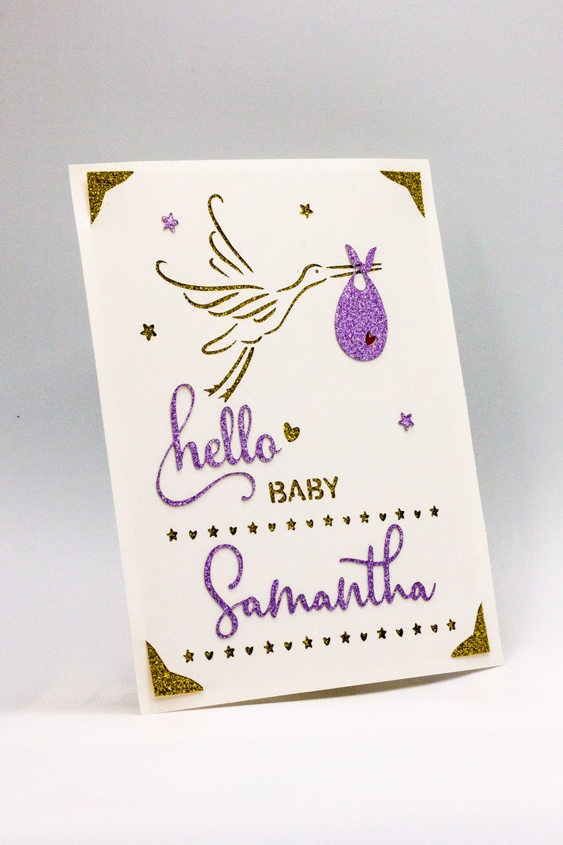 May include: A purple and gold glitter card featuring a stork carrying a baby in a purple and gold glitter baby bib. The card reads "hello baby Samantha" with gold glitter stars and hearts.
