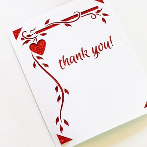 K&ouml;nnte beinhalten: Eine wei&szlig;e Karte mit einem roten Glitzerdesign aus einer Rebe mit Bl&auml;ttern und einem Herzen. Die Karte sagt "Danke!" in rotem Glitzer.