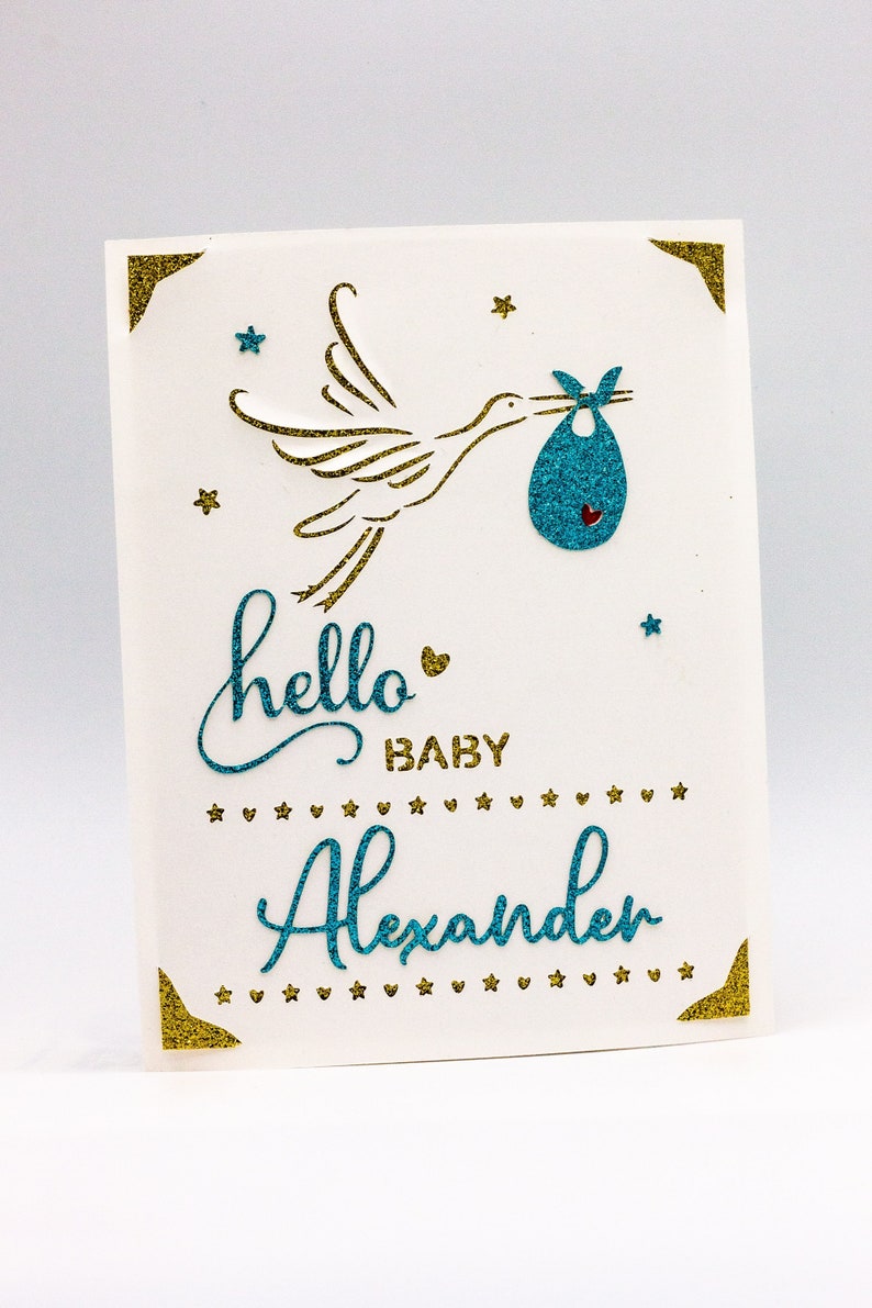 May include: A white card with gold glitter accents and a blue glitter stork carrying a blue glitter baby bundle. The card says "hello baby Alexander" in blue glitter.