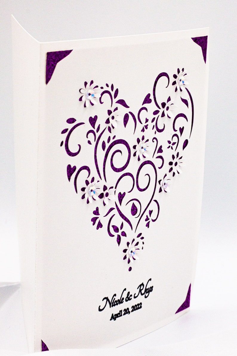 May include: A white card with a purple heart-shaped design featuring intricate swirls and flowers. The text "Nicole & Rhys" and "April 20, 2022" is printed in black at the bottom of the card.