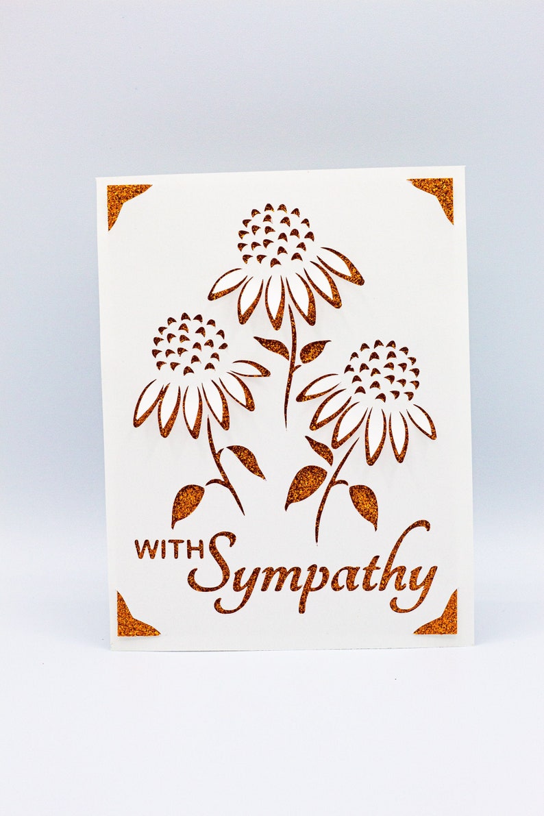 Puede incluir: Una tarjeta blanca con un borde de purpurina dorada y un dise&ntilde;o recortado de tres flores. El texto "With Sympathy" est&aacute; escrito en purpurina dorada.