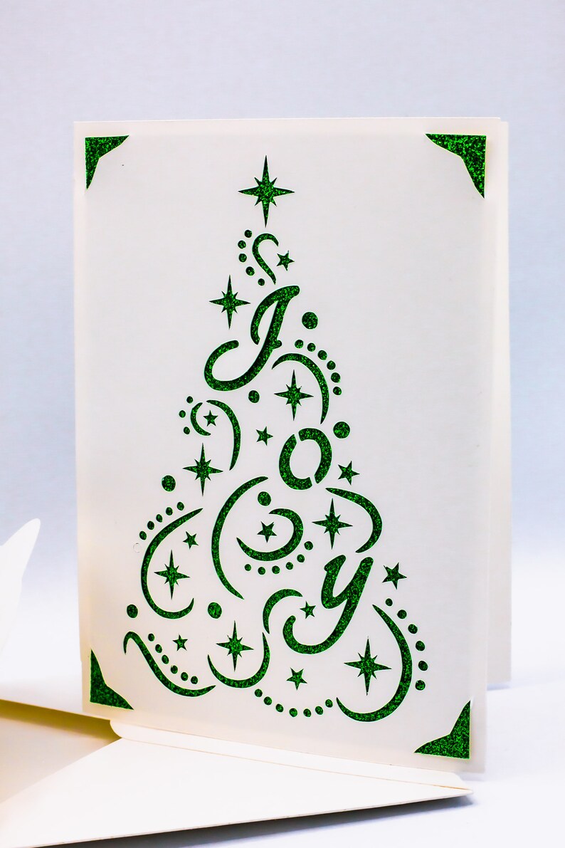 May include: A white card with a green glitter design of a Christmas tree with the word "Joy" inside. The tree is decorated with stars and swirls.