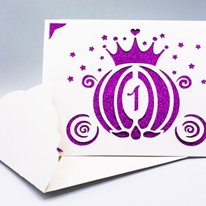 May include: A white card with a purple glitter design of a carriage with a crown on top. The number "1" is in the center of the carriage. The card has a scalloped edge.