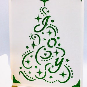 May include: A white card with a green glitter design of a Christmas tree with the word "Joy" inside. The tree is decorated with stars and swirls.