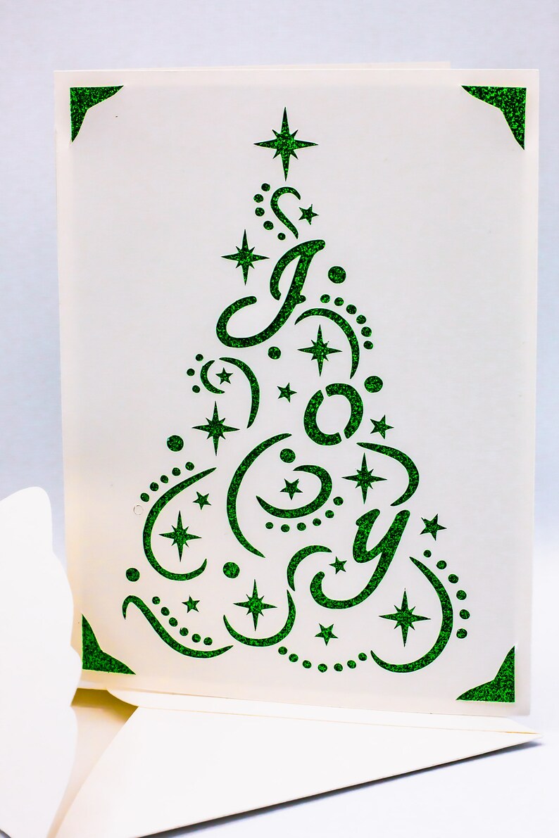 May include: A white card with a green glitter design of a Christmas tree with the word "Joy" inside. The tree is decorated with stars and swirls.