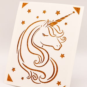Puede incluir: Una tarjeta blanca con un dise&ntilde;o de unicornio de purpurina dorada. El unicornio tiene un ojo cerrado y un cuerno que se curva hacia la derecha. El fondo est&aacute; lleno de peque&ntilde;as estrellas dor&eacute;es de purpurina.