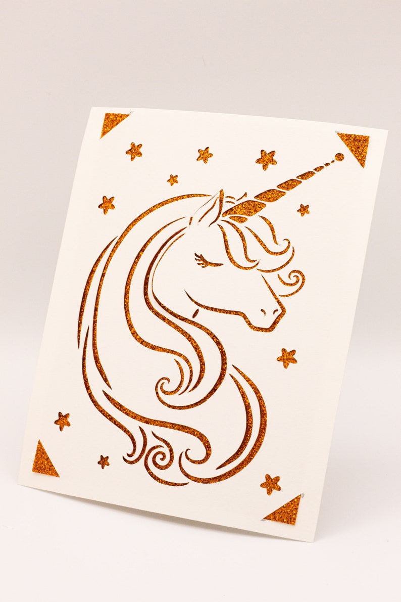 Puede incluir: Una tarjeta blanca con un dise&ntilde;o de unicornio de purpurina dorada. El unicornio tiene un ojo cerrado y un cuerno que se curva hacia la derecha. El fondo est&aacute; lleno de peque&ntilde;as estrellas dor&eacute;es de purpurina.