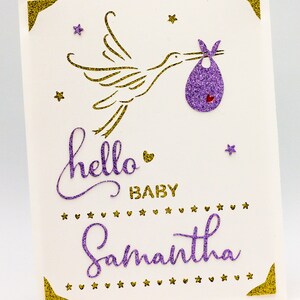 May include: A white card with gold glitter accents and purple glitter lettering. The card features a gold glitter stork carrying a purple glitter baby bib. The text on the card reads "hello baby Samantha".