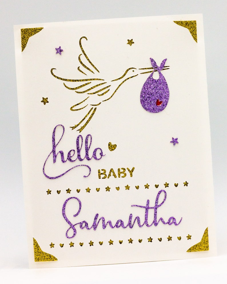 May include: A white card with gold glitter accents and purple glitter lettering. The card features a gold glitter stork carrying a purple glitter baby bib. The text on the card reads "hello baby Samantha".