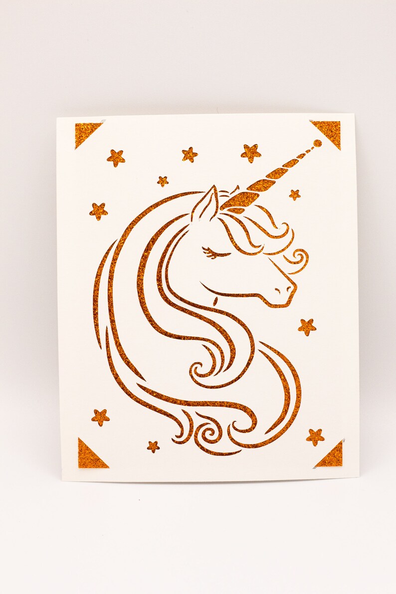 Puede incluir: Una tarjeta blanca con una silueta de unicornio dorada con purpurina y estrellas. El unicornio tiene un ojo cerrado y un cuerno rizado.