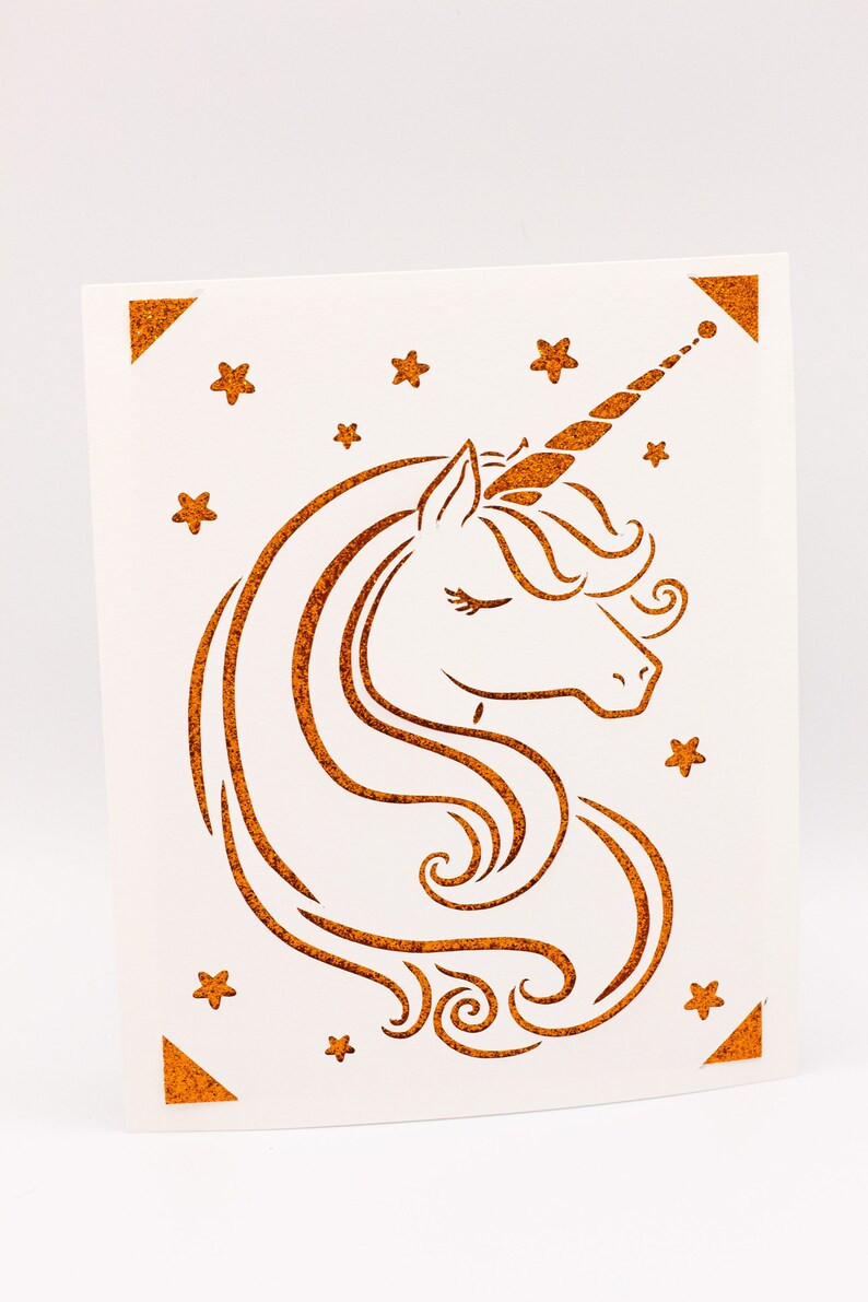 Puede incluir: Una tarjeta blanca con una silueta de unicornio marr&oacute;n con purpurina y estrellas. El unicornio tiene un ojo cerrado y un cuerno rizado.