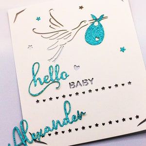 May include: A white card with a blue glitter stork carrying a baby in a blue glitter onesie. The card says "hello baby" and "Alexander" in blue glitter.