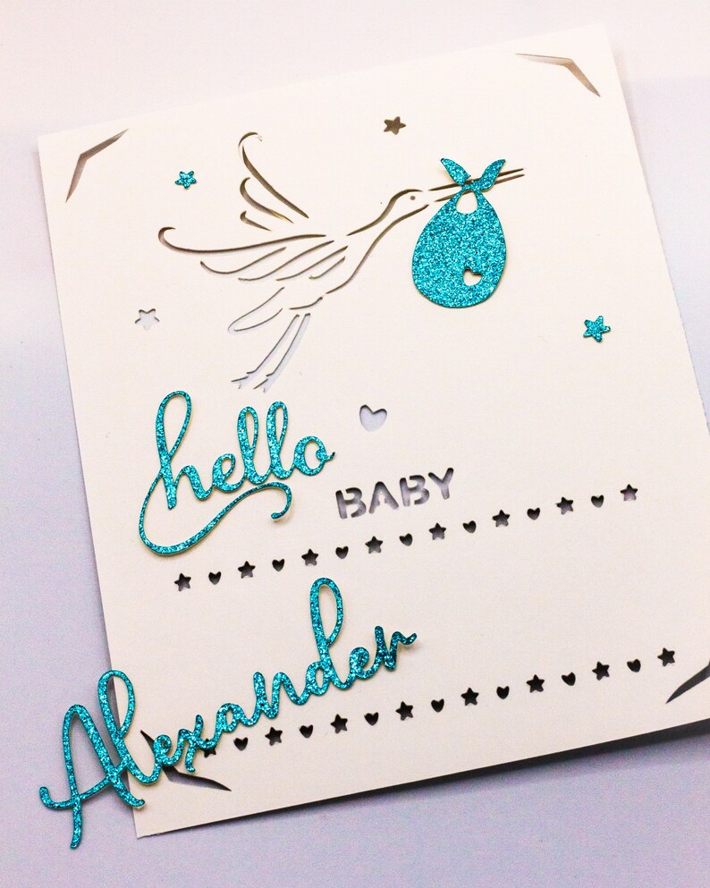 May include: A white card with a blue glitter stork carrying a baby in a blue glitter onesie. The card says "hello baby" and "Alexander" in blue glitter.