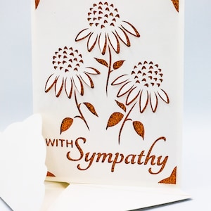 Puede incluir: Una tarjeta blanca con un borde de purpurina marr&oacute;n y un dise&ntilde;o recortado de tres flores. El texto "With Sympathy" est&aacute; escrito en purpurina marr&oacute;n en la tarjeta.