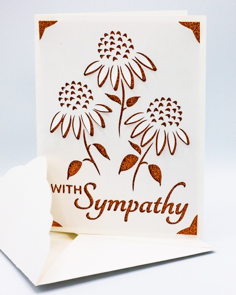 Puede incluir: Una tarjeta blanca con un borde de purpurina marr&oacute;n y un dise&ntilde;o recortado de tres flores. El texto "With Sympathy" est&aacute; escrito en purpurina marr&oacute;n en la tarjeta.