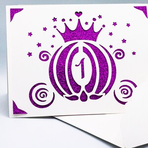 May include: A white card with a purple glitter design of a carriage with a crown on top. The carriage has a number "1" inside. The card is decorated with stars and swirls.