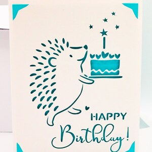 May include: A white card with a teal blue hedgehog holding a teal blue cake with a teal blue star on top. The card says "Happy Birthday!" in teal blue lettering.