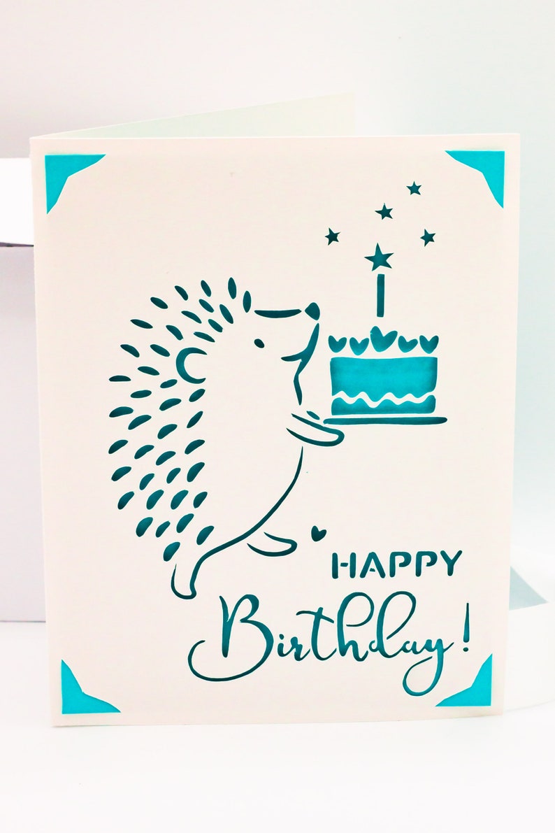 May include: A white card with a teal blue hedgehog holding a teal blue cake with a teal blue star on top. The card says "Happy Birthday!" in teal blue lettering.