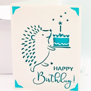 May include: A white card with a teal blue border and a hedgehog holding a teal blue cake with a single candle. The card says "Happy Birthday!" in teal blue lettering.
