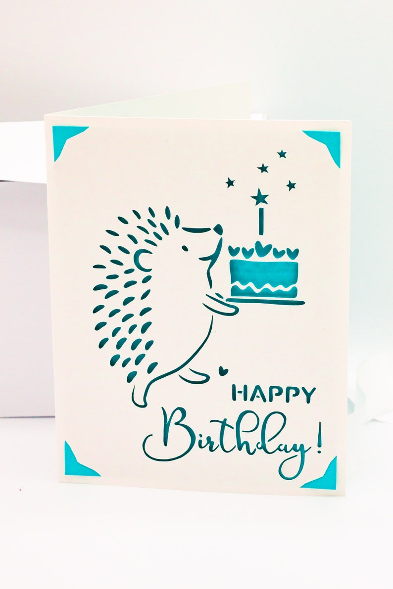 May include: A white card with a teal blue border and a hedgehog holding a teal blue cake with a single candle. The card says "Happy Birthday!" in teal blue lettering.