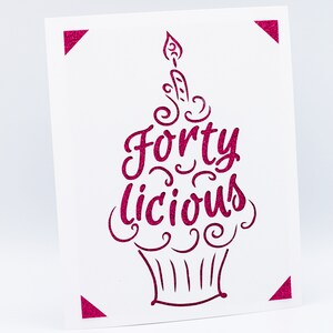 May include: A white card with a pink cupcake design. The cupcake has a candle on top and the words "Forty Licious" written on it.