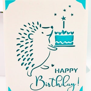 May include: A white card with a teal blue hedgehog holding a teal blue cake with a single candle and stars. The card says "Happy Birthday!" in teal blue.