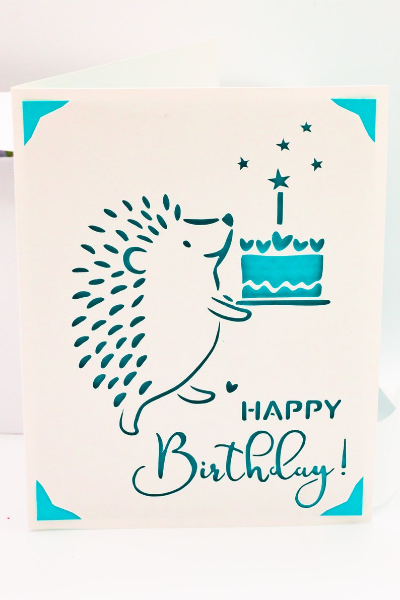 May include: A white card with a teal blue hedgehog holding a teal blue cake with a single candle and stars. The card says "Happy Birthday!" in teal blue.