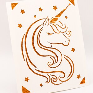 Puede incluir: Una tarjeta blanca con una silueta de unicornio dorada con purpurina y estrellas. El unicornio tiene un ojo cerrado y un cuerno rizado.
