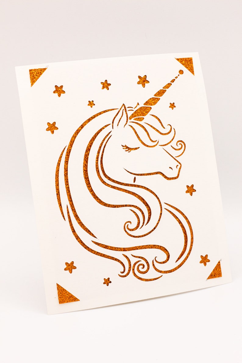 Puede incluir: Una tarjeta blanca con una silueta de unicornio dorada con purpurina y estrellas. El unicornio tiene un ojo cerrado y un cuerno rizado.