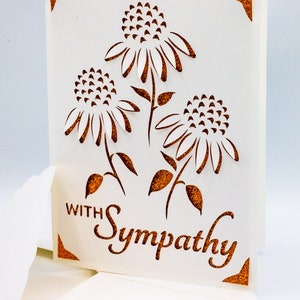 Puede incluir: Una tarjeta blanca con un dise&ntilde;o recortado de tres flores con detalles de purpurina marr&oacute;n. La tarjeta dice "Con simpat&iacute;a" en purpurina marr&oacute;n.
