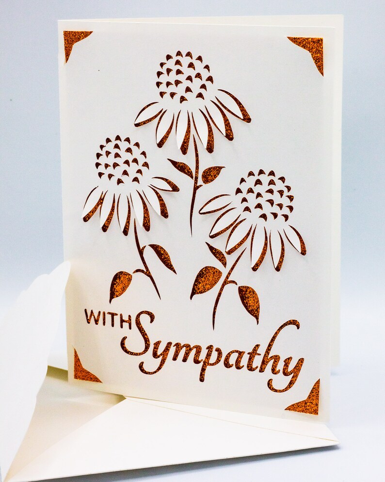 Puede incluir: Una tarjeta blanca con un dise&ntilde;o recortado de tres flores con detalles de purpurina marr&oacute;n. La tarjeta dice "Con simpat&iacute;a" en purpurina marr&oacute;n.