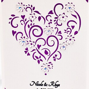 May include: A white card with a purple glitter border and a purple heart-shaped design with white flowers and rhinestones. The text "Nicole & Rhys" and "April 20, 2022" is printed below the design.