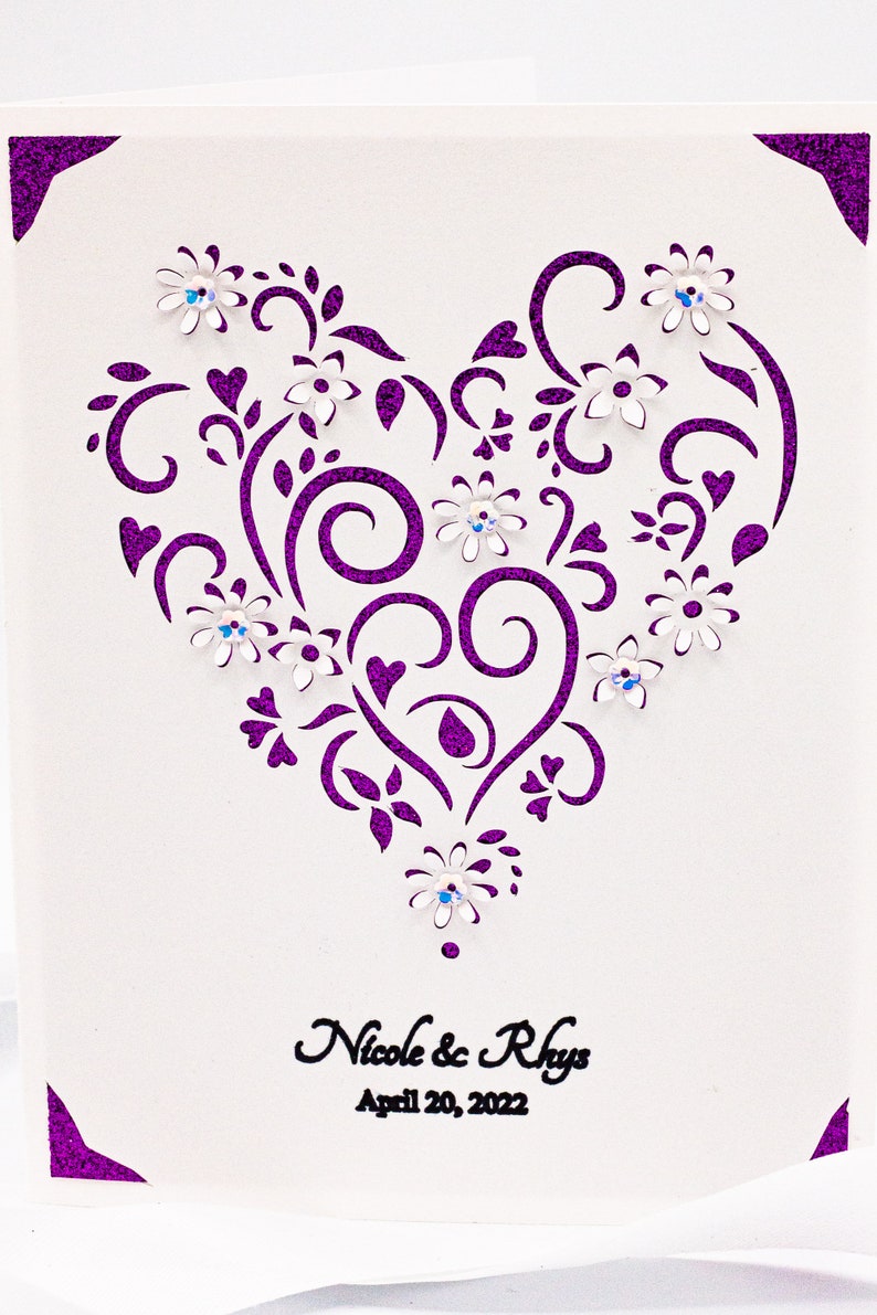 May include: A white card with a purple glitter border and a purple heart-shaped design with white flowers and rhinestones. The text "Nicole & Rhys" and "April 20, 2022" is printed below the design.