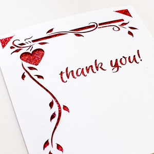 K&ouml;nnte beinhalten: Eine wei&szlig;e Karte mit einem roten Glitzer-Design aus einer Rebe mit Bl&auml;ttern und einem Herzen. Die Karte sagt "Danke!" in rotem Glitzer.