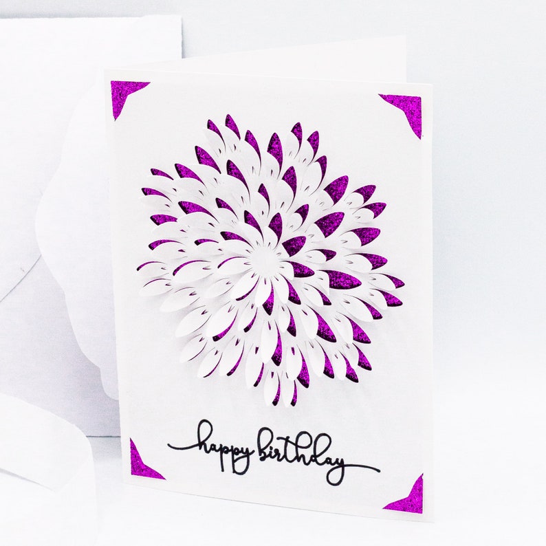 May include: A white card with a purple glitter border and a white paper flower in the center. The card says "happy birthday" in black script.