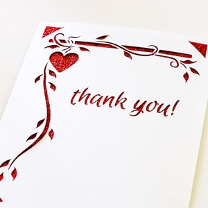 K&ouml;nnte beinhalten: Eine wei&szlig;e Karte mit einem roten Glitzerdesign aus einer Rebe mit Bl&auml;ttern und einem Herzen. Die Karte sagt "Danke!" in rotem Glitzer.