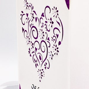 May include: A white card with a purple heart-shaped design featuring intricate swirls and flowers. The card is decorated with small white flowers and has the text "Nicole & Rhys" and "April 20, 2022" printed in black.