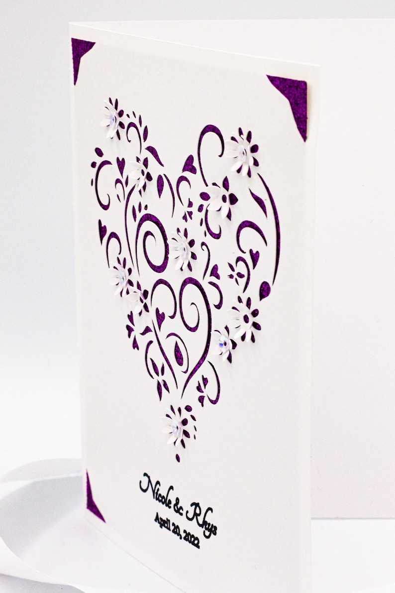May include: A white card with a purple heart-shaped design featuring intricate swirls and flowers. The card is decorated with small white flowers and has the text "Nicole & Rhys" and "April 20, 2022" printed in black.