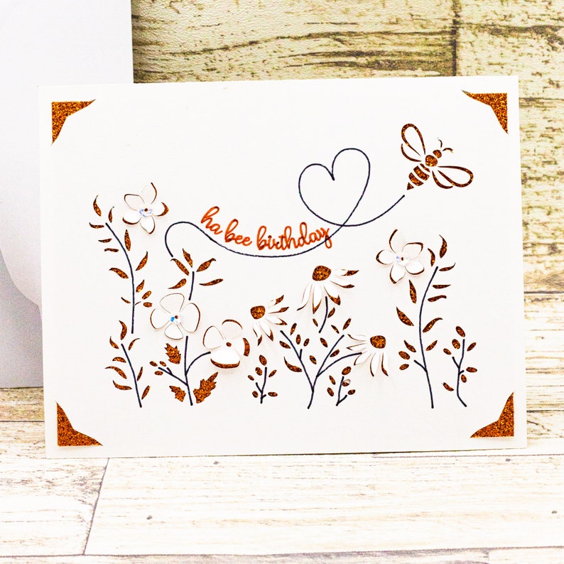 May include: White card with a bee and flowers cut out. The card has a copper coloured border and the text "ha bee birthday" is written in copper coloured lettering.