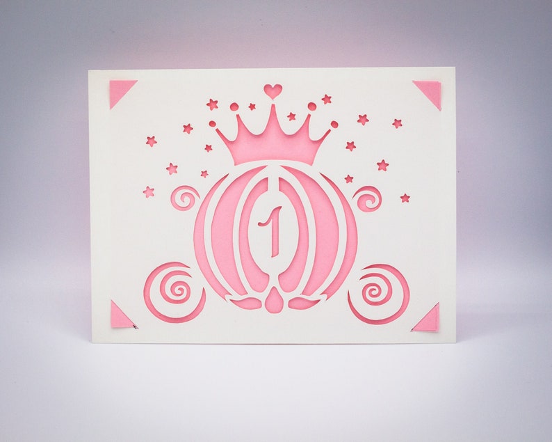 May include: White card with a pink princess carriage cut out design. The carriage has a pink crown with a heart on top and the number 1 inside. There are small stars around the carriage.