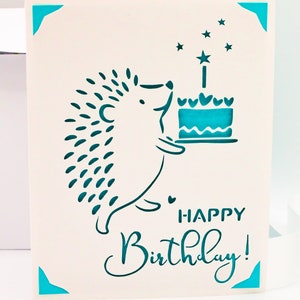 May include: A white card with a teal blue hedgehog holding a teal blue cake with a single candle and stars. The card says "Happy Birthday!" in teal blue lettering.